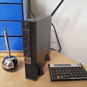 May include: A black Dell OptiPlex 7040 computer tower with a silver pen in a chrome pen holder and a vintage calculator on a wooden desk. The computer has a black antenna and the Dell logo.