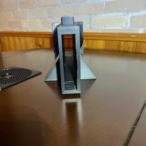 Peut inclure: Un objet noir imprim&eacute; en 3D avec un cadre rectangulaire et une base triangulaire. L'objet est pos&eacute; sur une surface marron fonc&eacute;, avec un mur de briques en arri&egrave;re-plan. Un composant circulaire perfor&eacute; est visible &agrave; gauche.
