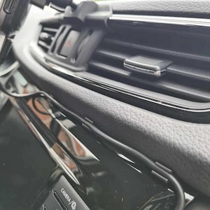 Könnte beinhalten: Nahaufnahme des Innenraums eines Autos mit einer schwarzen Handyhalterung, die an der Lüftung befestigt ist. Ein schwarzes Kabel ist mit der Handyhalterung verbunden und verläuft entlang des Armaturenbretts. Das Armaturenbrett hat eine schwarze und silberne Oberfläche. Die Radiobedienelemente des Autos sind in der unteren rechten Ecke des Bildes sichtbar. Die Radiobedienelemente haben einen schwarzen Hintergrund mit silbernen Knöpfen und der Text "CAMERA/GP" ist auf einem der Knöpfe sichtbar.