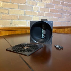 Peut inclure: Un composant &eacute;lectronique noir de forme carr&eacute;e avec une ouverture circulaire, un couvercle perfor&eacute; d&eacute;tach&eacute; et plusieurs vis. Le composant est ouvert, r&eacute;v&eacute;lant des circuits internes. Les &eacute;l&eacute;ments sont sur une surface en bois brun fonc&eacute;.