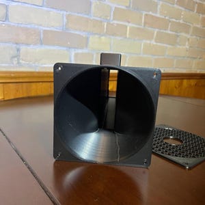Peut inclure: Un pavillon de haut-parleur noir imprim&eacute; en 3D, avec une face carr&eacute;e et une ouverture circulaire. Une plaque perfor&eacute;e noire s&eacute;par&eacute;e est pos&eacute;e sur le c&ocirc;t&eacute;. Le pavillon est sur une surface en bois brun, avec un mur de briques en arri&egrave;re-plan.