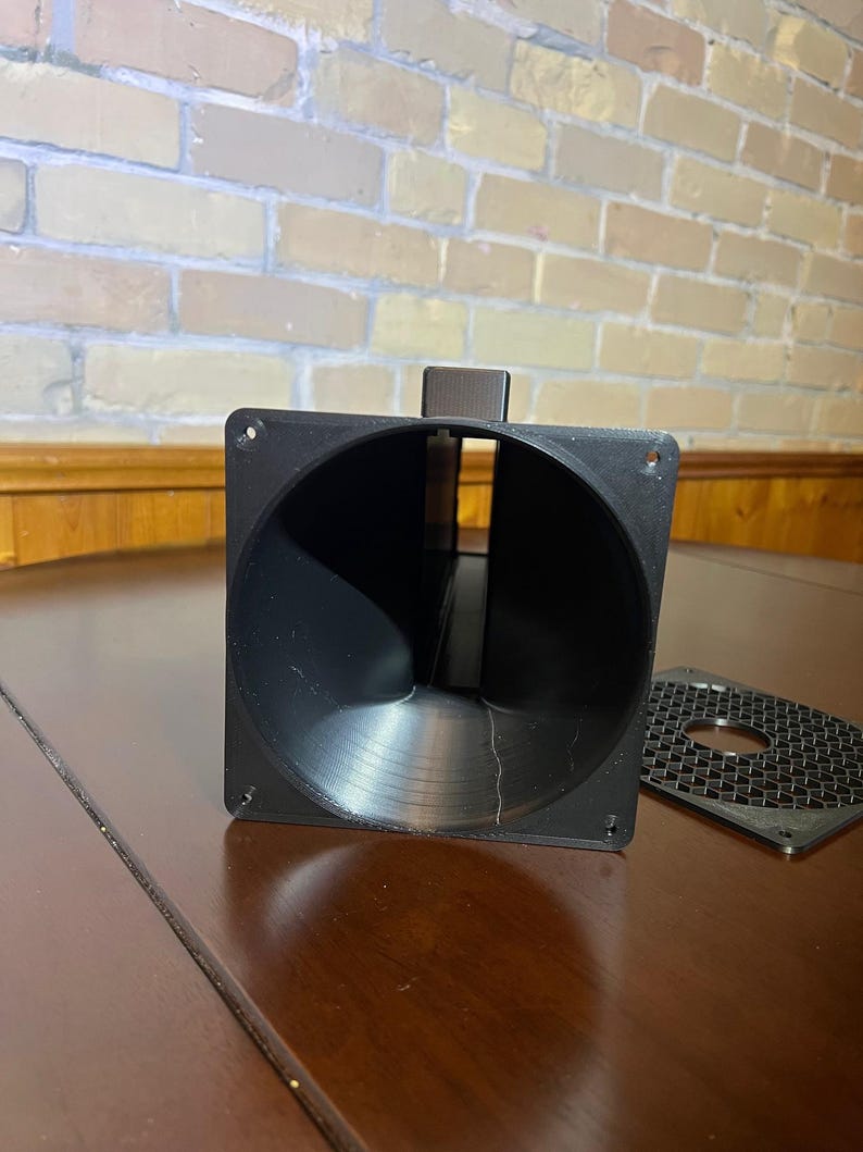 Peut inclure: Un pavillon de haut-parleur noir imprim&eacute; en 3D, avec une face carr&eacute;e et une ouverture circulaire. Une plaque perfor&eacute;e noire s&eacute;par&eacute;e est pos&eacute;e sur le c&ocirc;t&eacute;. Le pavillon est sur une surface en bois brun, avec un mur de briques en arri&egrave;re-plan.
