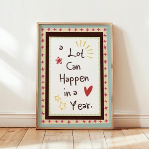 May include: A framed print with the text "A Lot Can Happen in a Year." The artwork features a white background with black text, a red heart, and decorative stars and flowers. The frame is light brown with a decorative border.