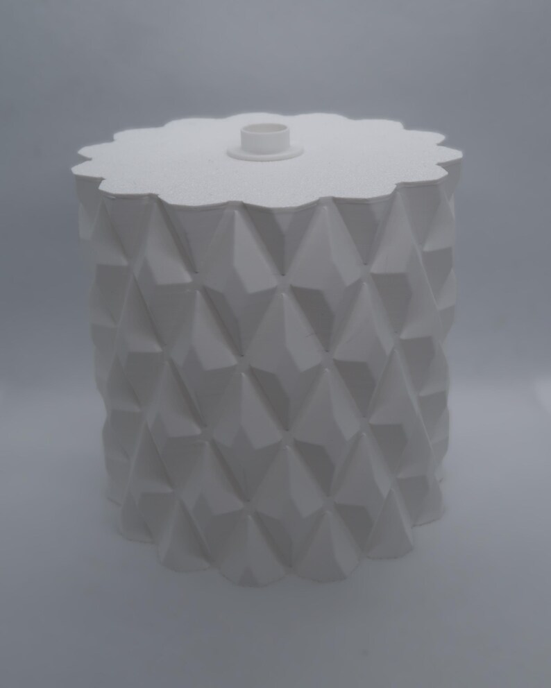 May include: A white, geometric-patterned container with a lid. The container has a cylindrical body with a repeating diamond pattern. The lid is scalloped and has a small, circular handle. The overall design is modern and minimalist.