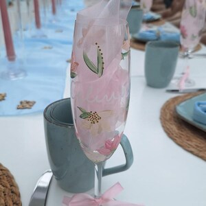 May include: A champagne flute with floral designs and a pink ribbon tied around the stem. The glass is filled with pink tissue paper. A blue mug and a silver knife are also visible.