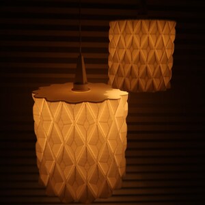 May include: Two pendant lamps with geometric patterned shades. The lampshades are cylindrical with a repeating diamond pattern. The lamps are lit, emitting a warm, soft glow. The lamps are suspended from the ceiling.
