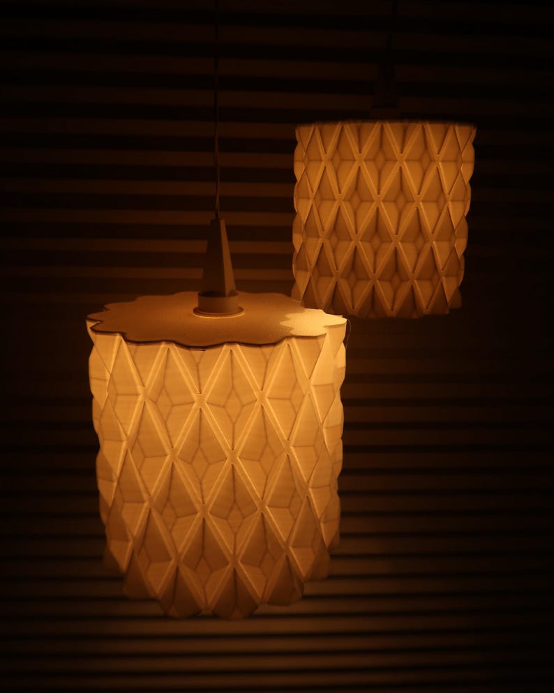May include: Two pendant lamps with geometric patterned shades. The lampshades are cylindrical with a repeating diamond pattern. The lamps are lit, emitting a warm, soft glow. The lamps are suspended from the ceiling.