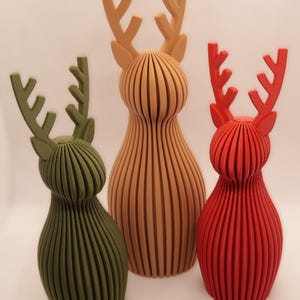 May include: Three decorative reindeer figurines in shades of green, tan, and red. Each reindeer has a ribbed body and stylized antlers. The figurines are arranged against a white background, showcasing their unique colors and designs.