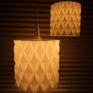 May include: Two geometric pendant lights with a warm glow. The lampshades have a repeating diamond pattern, creating a textured surface. The lights hang from the ceiling against a backdrop of horizontal lines.