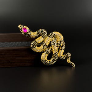 Puede incluir: Broche de serpiente dorada con detalles en negro y blanco. La cabeza de la serpiente presenta un ojo de gema rosa. El cuerpo está adornado con piedras negras y blancas, creando un contraste llamativo con la base dorada. Accesorio decorativo.