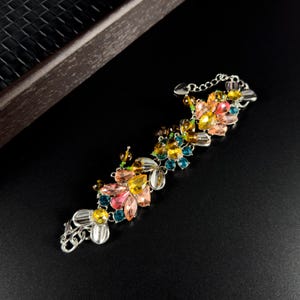 Op de afbeelding: Een zilverkleurige armband met een bloemmotief en kleurrijke edelstenen. De armband heeft roze, gele, blauwe en groene edelstenen in een cluster. Het heeft een ketting sluiting. De armband is ongeveer 18 cm lang.