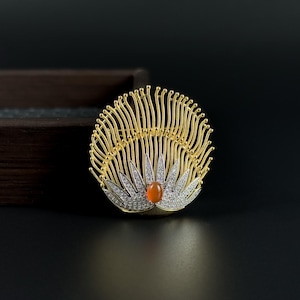 Op de afbeelding: Goudkleurige broche met een waaiervormig ontwerp. De broche heeft een centrale oranje edelsteen, zilverkleurige accenten en fijne gouddraad details. De achtergrond is donker.