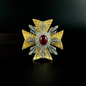 May include: A gold-toned cross-shaped brooch with a central oval-shaped, deep red gemstone. The brooch features a detailed design with diamond-like accents and a textured gold finish, set against a dark background.
