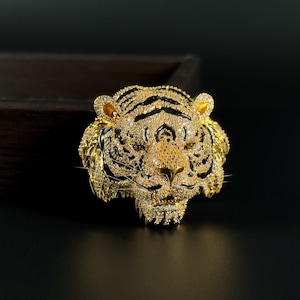May include: A gold-colored tiger head pendant with black stripes and sparkling crystal accents. The pendant is intricately detailed, with a textured surface and a fierce expression. The jewelry piece is set against a dark background.