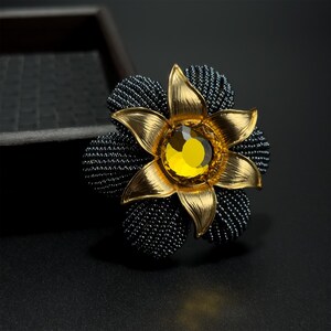 May include: A flower-shaped brooch with a gold-coloured metal design. The brooch features a central yellow gemstone and is surrounded by black beaded accents. The petals are detailed with a textured finish, creating a striking contrast.
