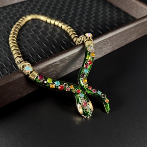 May include: A gold-coloured snake necklace featuring a green enamel body and colourful gemstone embellishments. The snake's body is decorated with a variety of colourful gems, including red, blue, and yellow. The necklace is set against a dark background.