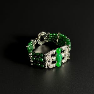 Könnte beinhalten: Ein grünes Perlenarmband mit silbernem Verschluss und Akzenten. Das Armband hat drei Reihen kleiner, runder grüner Perlen und ein rechteckiges silbernes Mittelstück mit klaren Steinen und drei größeren grünen Steinen.