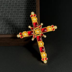May include: A gold-toned cross pendant featuring red and yellow gemstones. The cross has a central square with a pearl-like stone, surrounded by red rectangular stones and smaller yellow stones. The edges are adorned with red and clear accent stones.