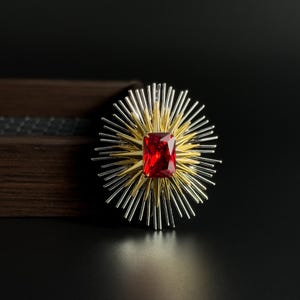Puede incluir: Broche llamativo con una gema roja rectangular en el centro, rodeada de púas doradas y plateadas. El diseño del broche destaca sobre un fondo oscuro, resaltando sus intrincados detalles y colores vibrantes. Un accesorio llamativo.