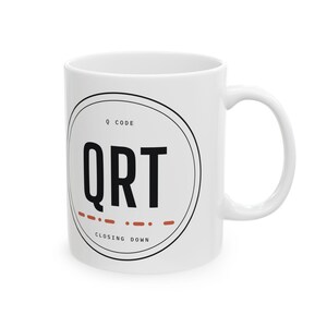 May include: A white ceramic mug featuring a circular black graphic with the text Q CODE, QRT, and CLOSING DOWN. The design includes a series of orange dashes arranged in a line below the letters QRT, centred on the front of the mug.