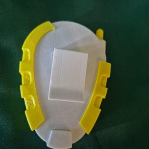 May include: A 3D-printed object with a white oval base, a rectangular white centre, and yellow curved side pieces. The object has a small, rounded yellow piece on top and a white rectangular piece at the bottom.