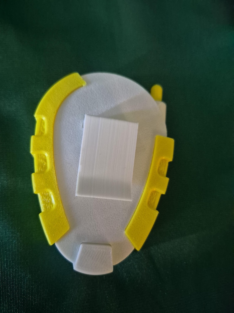 May include: A 3D-printed object with a white oval base, a rectangular white centre, and yellow curved side pieces. The object has a small, rounded yellow piece on top and a white rectangular piece at the bottom.