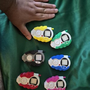 May include: Six colourful handheld digital devices in various colours: yellow, green, black, blue, red, and pink. Each device has a white base, a small screen, and buttons. The devices are on a green surface.