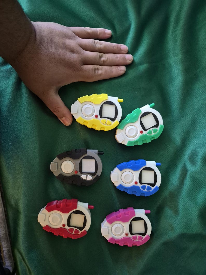 May include: Six colourful handheld digital devices in various colours: yellow, green, black, blue, red, and pink. Each device has a white base, a small screen, and buttons. The devices are on a green surface.