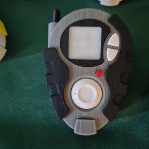 May include: A grey and black handheld device featuring a small screen, white buttons, and a red button. The device has a circular white button at the bottom and a small antenna. Other similar devices are visible in the background.