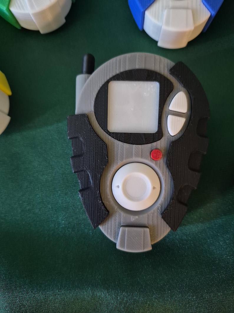 May include: A grey and black handheld device featuring a small screen, white buttons, and a red button. The device has a circular white button at the bottom and a small antenna. Other similar devices are visible in the background.
