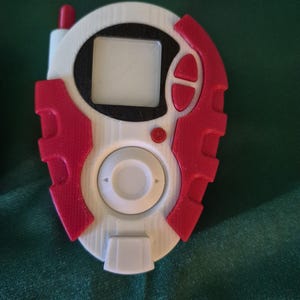 May include: A white and red handheld device with a small screen and buttons. The device has a circular centre with a rectangular screen and a red antenna. The device is likely a toy or collectible item.