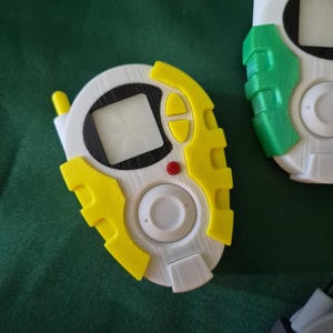 May include: Close-up of several white, yellow, and green handheld digital devices. The devices have a screen, buttons, and an antenna. The devices are on a green surface.