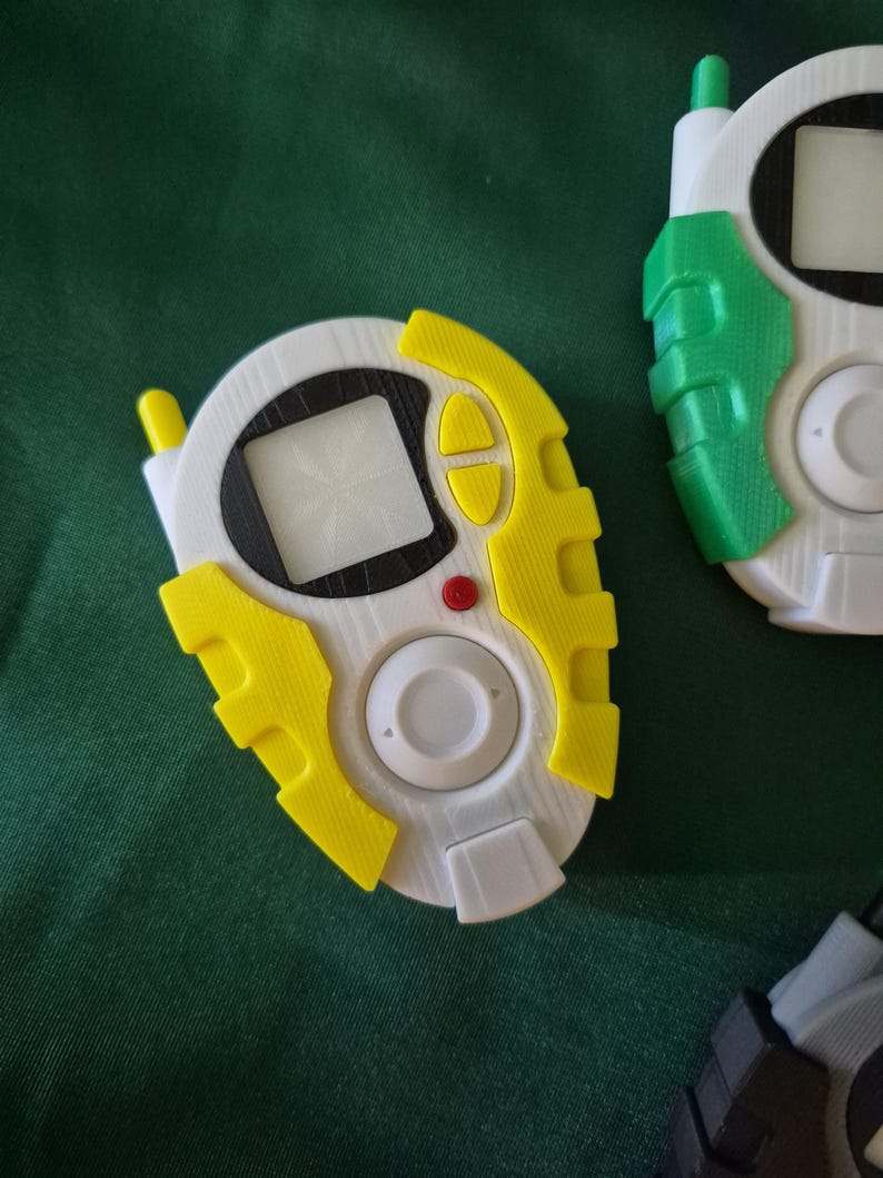 May include: Close-up of several white, yellow, and green handheld digital devices. The devices have a screen, buttons, and an antenna. The devices are on a green surface.