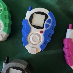 May include: A selection of 3D-printed digital pet devices in various colours. The central device is white with blue detailing, a small screen, and a red button. Other devices are green, grey, and pink.