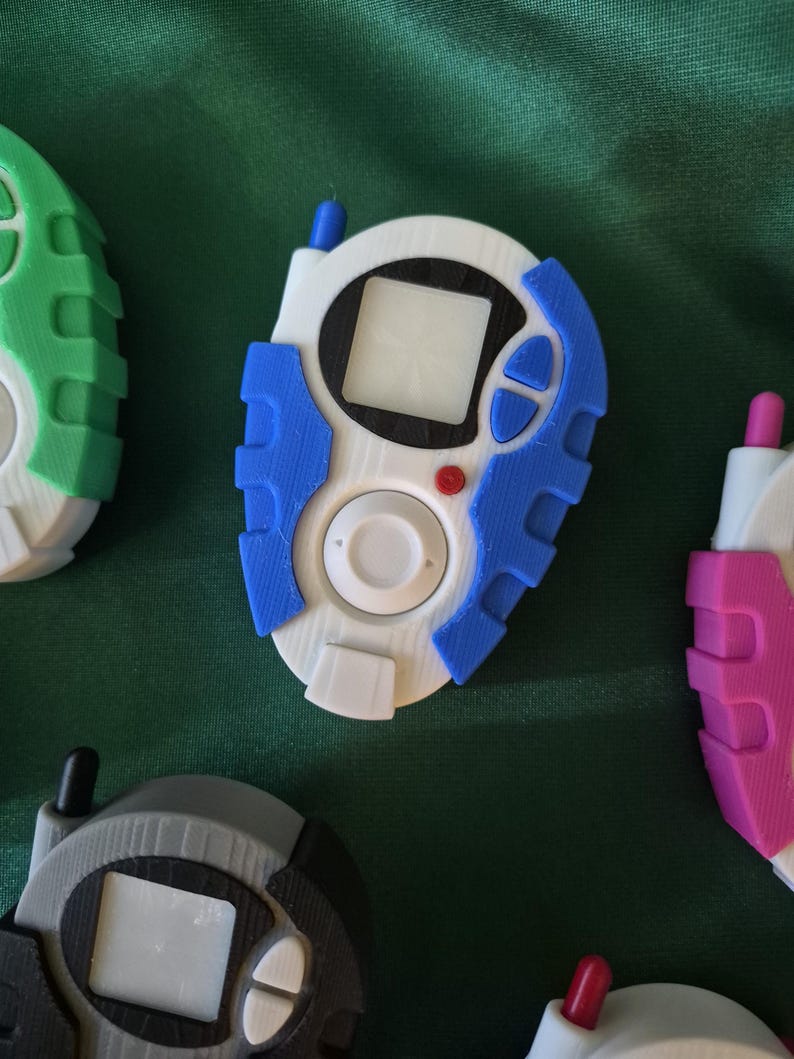 May include: A selection of 3D-printed digital pet devices in various colours. The central device is white with blue detailing, a small screen, and a red button. Other devices are green, grey, and pink.