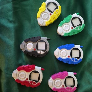 May include: Six handheld digital devices in various colours, including yellow, green, blue, red, pink, and grey. Each device has a white screen, a button, and a small antenna. The background is a dark green fabric.