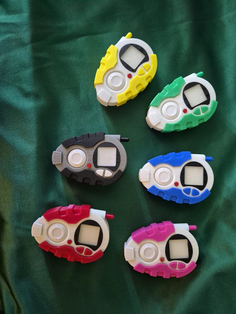 May include: Six handheld digital devices in various colours, including yellow, green, blue, red, pink, and grey. Each device has a white screen, a button, and a small antenna. The background is a dark green fabric.