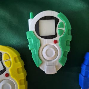 May include: A white, green, and black handheld device with a small screen, buttons, and a central dial. The device has a green antenna and accents. Other similar devices in yellow and blue are partially visible.