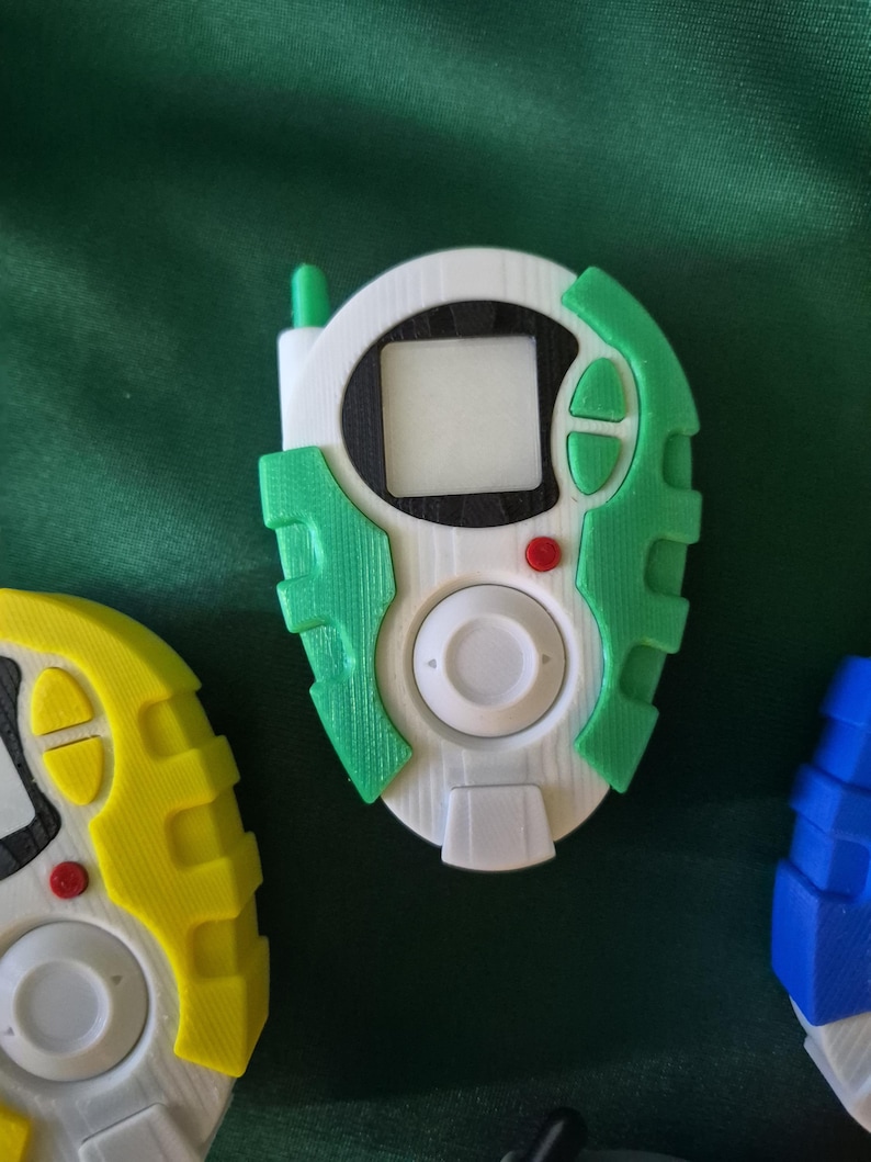 May include: A white, green, and black handheld device with a small screen, buttons, and a central dial. The device has a green antenna and accents. Other similar devices in yellow and blue are partially visible.