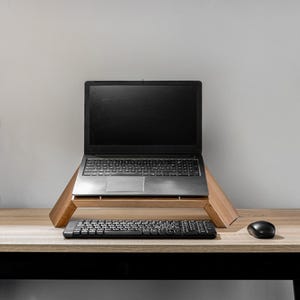 Op de afbeelding: Een zwarte laptop op een houten standaard met een zwart toetsenbord en muis op een lichtbruin houten bureau.