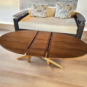 Op de afbeelding: Een ovale houten salontafel met een donkerbruine afwerking en een centraal tussenstuk. De tafel heeft een lichtgekleurd houten onderstel met schuine poten en staat voor een bank met donkerbruine armleuningen en twee kussens met een patroon.
