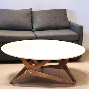 May include: A white oval coffee table with a wooden base featuring angled legs and a dark gray two-cushion sofa positioned directly behind it. The table has a smooth, solid white top supported by a brown wooden frame with visible metal hardware.
