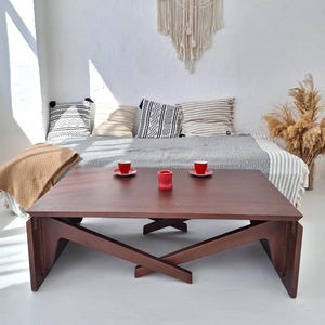 May include: A dark brown wooden coffee table with an X-shaped base. Two red cups and a red candle holder sit on the table. The table is in a white room with a bed, pillows, and a macrame wall hanging.