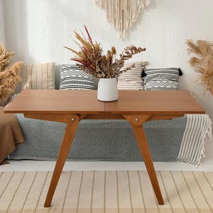 Può includere: Un tavolo rettangolare in legno con una finitura liscia color marrone medio e gambe angolate. Il tavolo presenta una giuntura centrale che indica un design pieghevole o estensibile, con un vaso bianco testurizzato contenente fiori secchi.