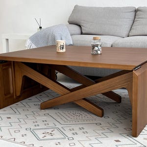 May include: A rectangular wooden coffee table with a smooth brown finish and a cross-legged base design. The table features a flat top surface with two small decorative items, including a white container with the black Japanese character for Zen.
