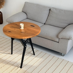 May include: A round wooden side table with a natural wood grain finish. The table has a black metal base and three angled legs. A small decorative cup sits on the table's surface. A gray sofa is in the background.