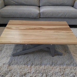 May include: A square wooden coffee table with a light brown tabletop and grey base. The table sits on a fluffy, light grey rug in front of a grey sofa. The wood grain is visible on the tabletop.