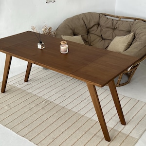 K&ouml;nnte beinhalten: Ein rechteckiger Holzesstisch mit glatter, dunkelbrauner Oberfl&auml;che und vier schr&auml;gen Beinen. Die Tischplatte weist eine sichtbare Naht auf, die auf ein ausziehbares Design hindeutet. Ein kleiner Glasdiffusor und eine Kerze stehen auf dem Tisch.