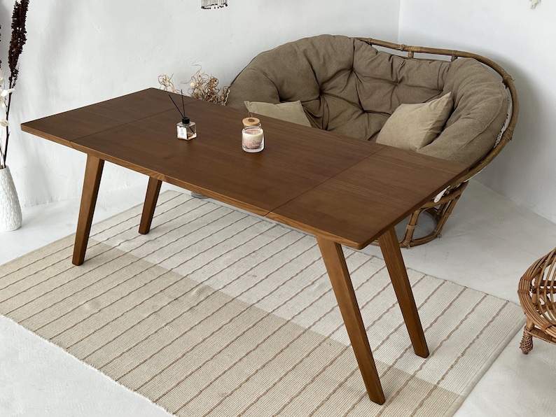K&ouml;nnte beinhalten: Ein rechteckiger Holzesstisch mit glatter, dunkelbrauner Oberfl&auml;che und vier schr&auml;gen Beinen. Die Tischplatte weist eine sichtbare Naht auf, die auf ein ausziehbares Design hindeutet. Ein kleiner Glasdiffusor und eine Kerze stehen auf dem Tisch.