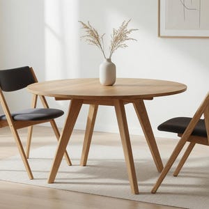 May include: A round light oak dining table with four angled legs and two matching chairs featuring dark gray upholstered seats and backrests. A white ceramic vase containing dried pampas grass sits in the center of the table surface.
